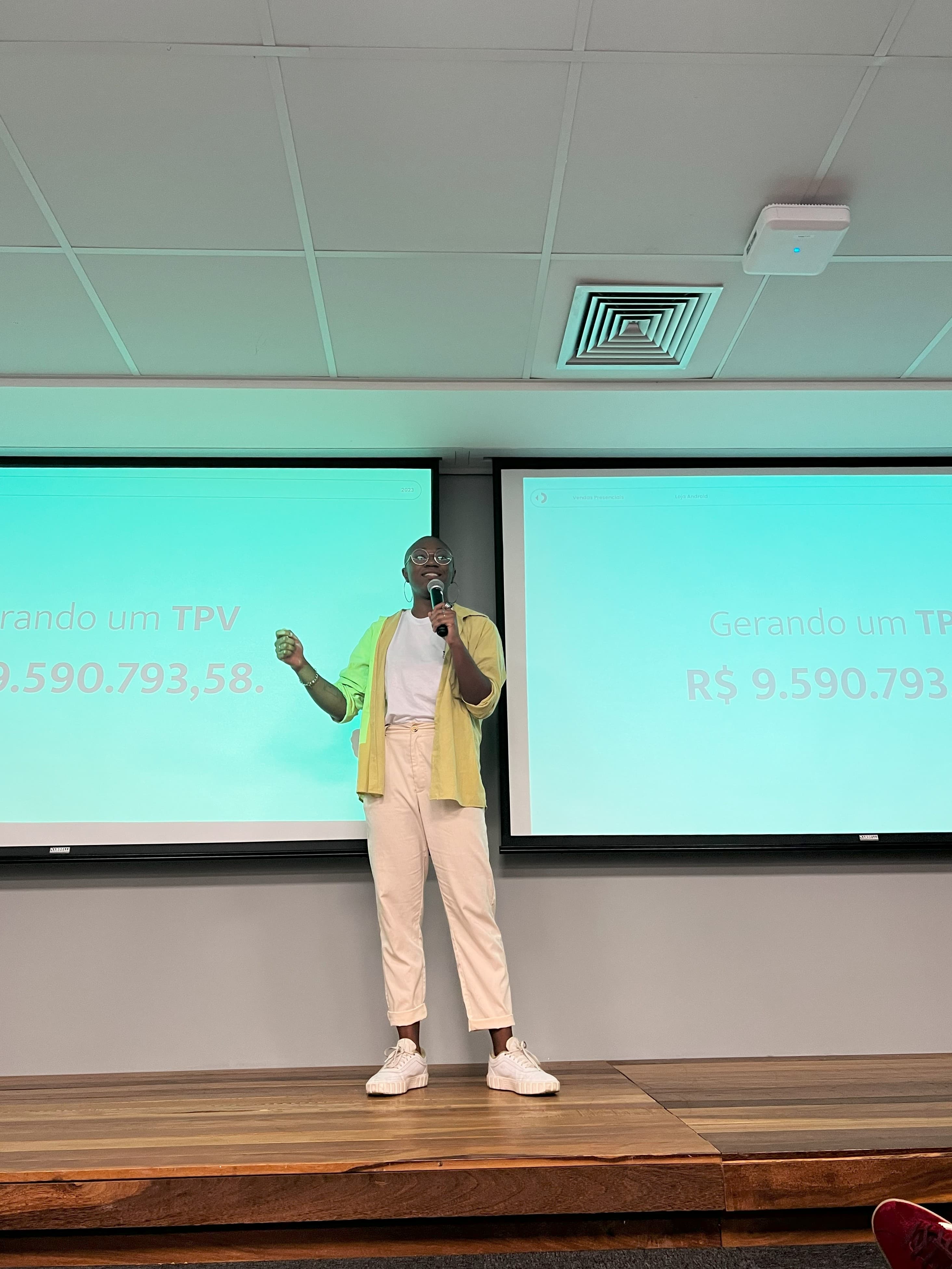Dandara standing on a stage in front of a projected green screen, she is holding a mic and mid-speech