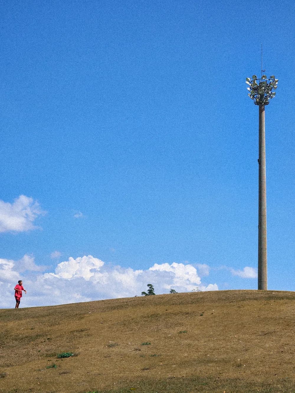 A boy in red at the far left edge of a dry grass hill, the floodlight pole standing at the right under a wide blue sky.
