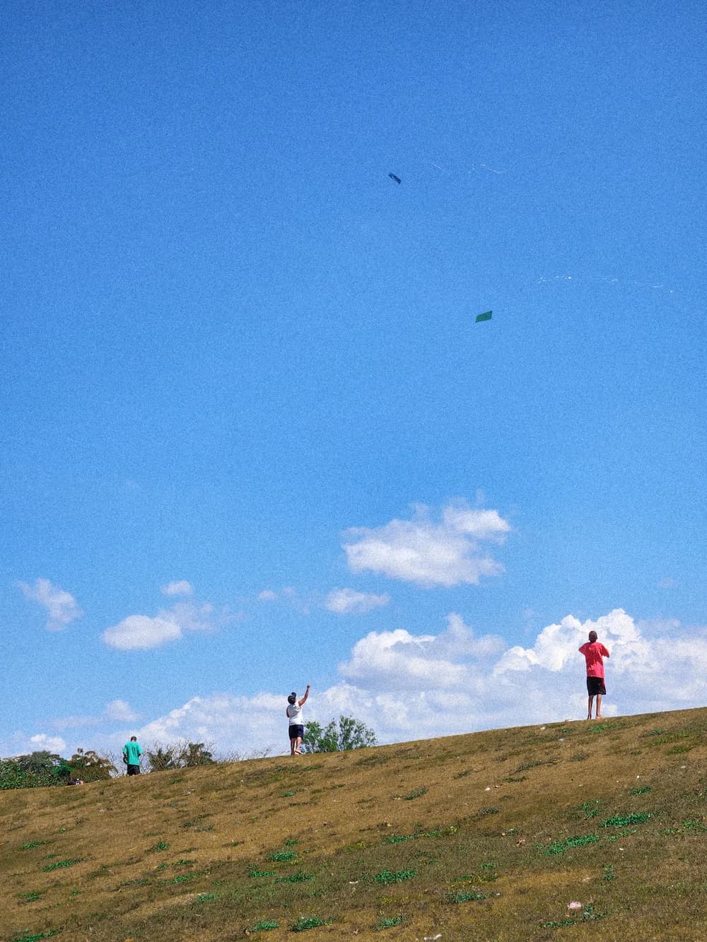 Three people on the crest of a grassy hill flying kites, two kites small against a blue sky with low clouds.