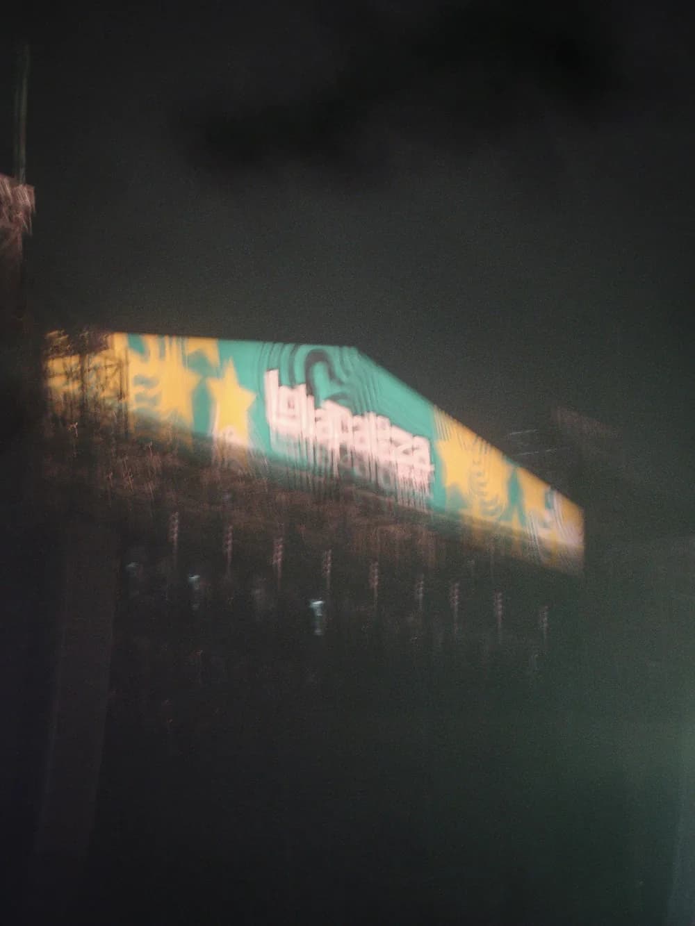 A blurred night view of the Lollapalooza sign, yellow and teal, glowing above a dark structure.