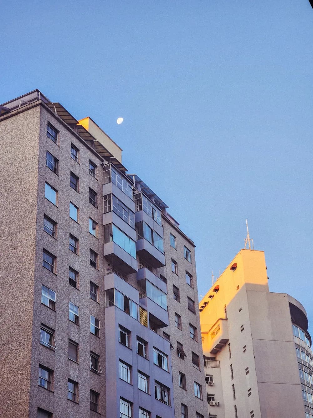 Apartment blocks at dusk, a small moon above the rooftops and the last orange light on a tower at the right.