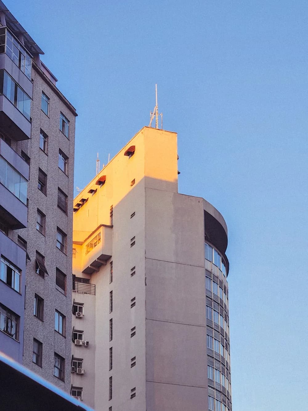 The top of a pale tower catching orange evening light beside a speckled apartment block, under a clear blue sky.