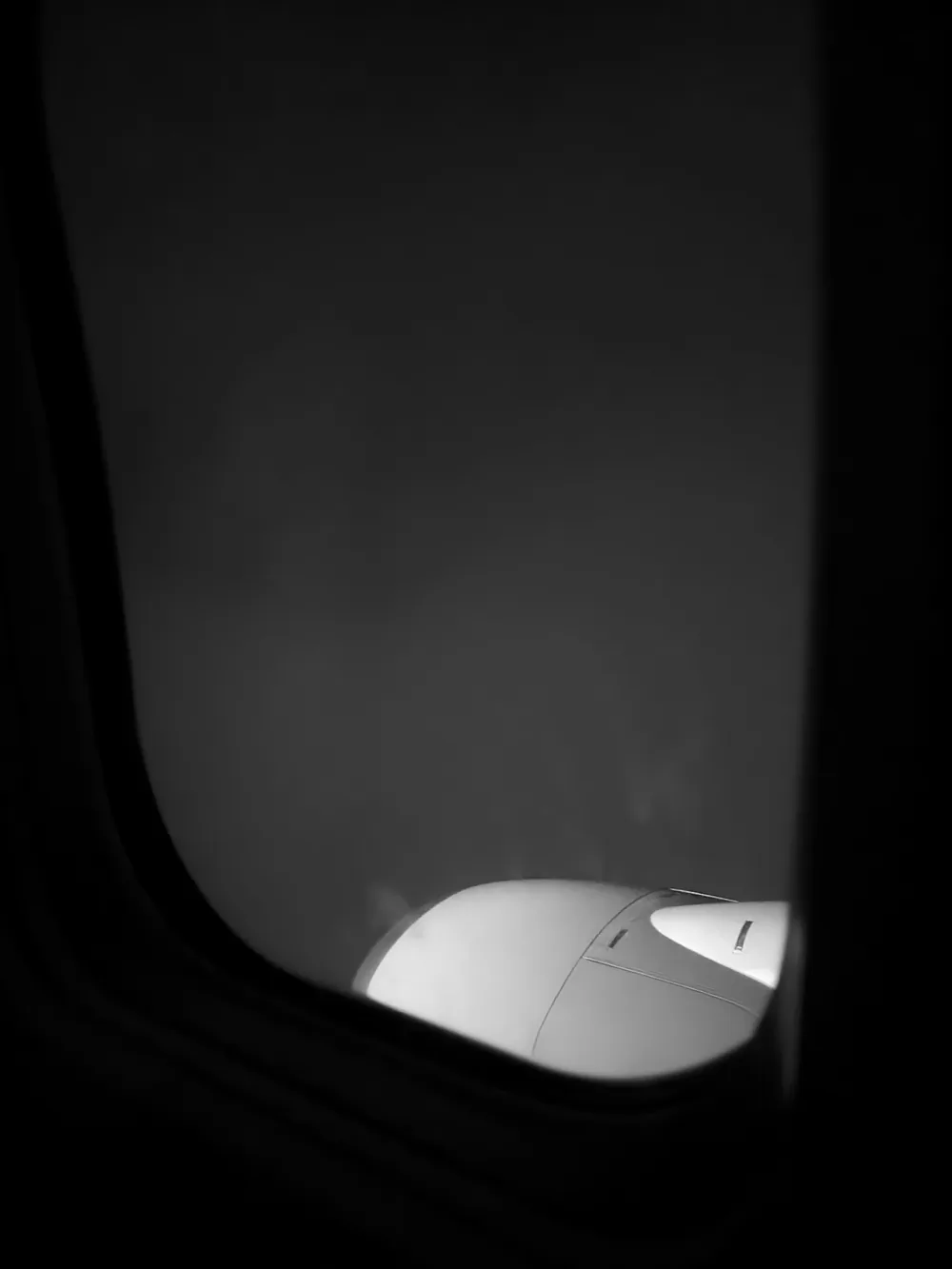 Black and white view through an aeroplane window into grey fog, the curve of an engine glowing at the bottom.
