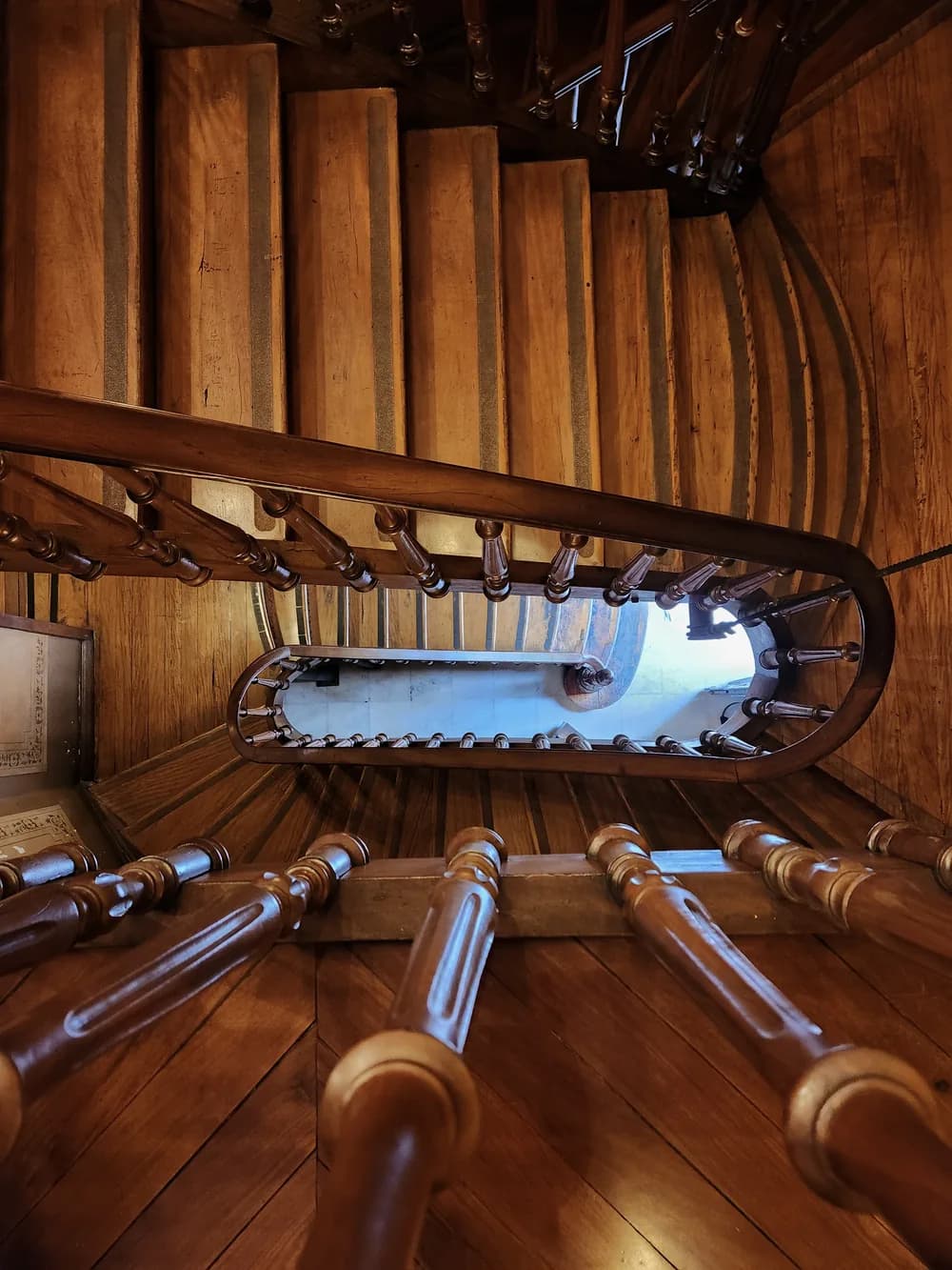 Looking down an oval wooden stairwell, turned balusters circling a pale floor far below.