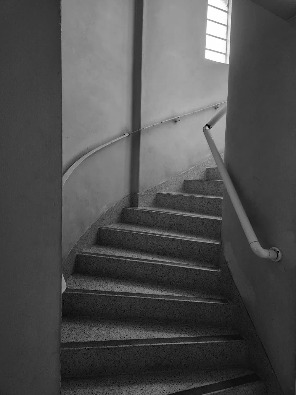 Black and white curving terrazzo staircase between plain walls, a handrail climbing towards a small bright window.