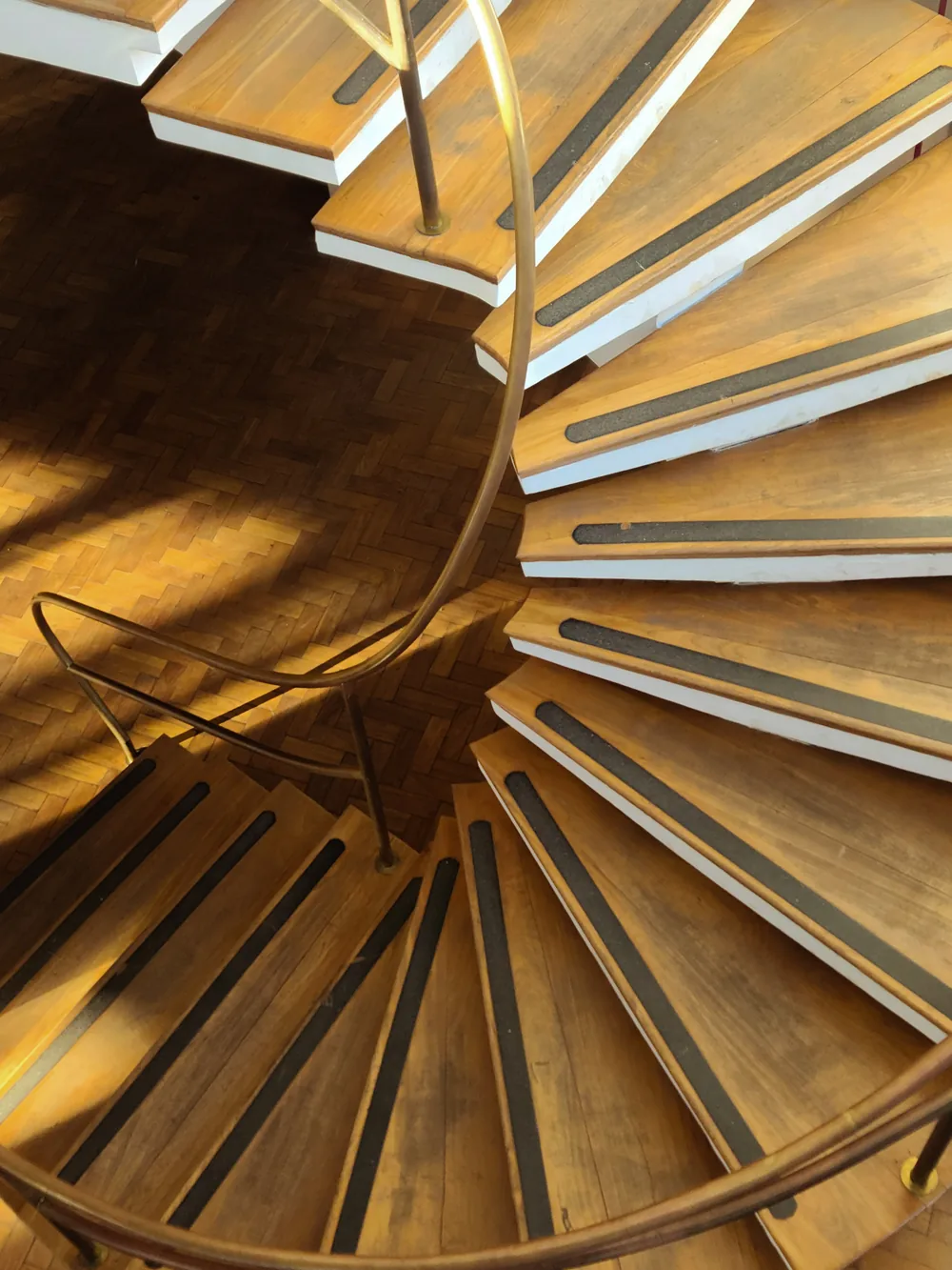 A wooden spiral staircase with a brass rail seen from above, sunlight across a herringbone floor.